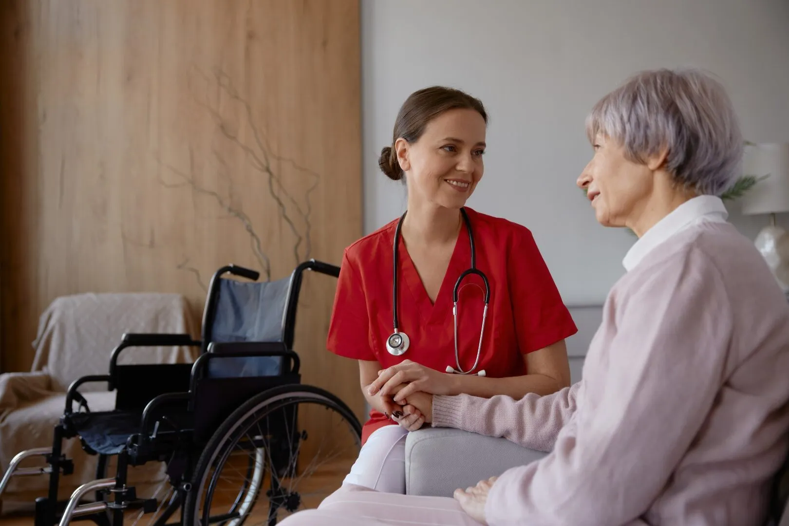 Female doctor talking to her patient during home visit. Woman medical practitioner supporting and helping elderly lady in wheelchair. Happy retirement with professional service